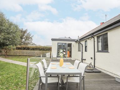 An outdoor dining table with six chairs on a wooden deck next to a house with light walls and dark window frames at Laneton in Rhosneigr