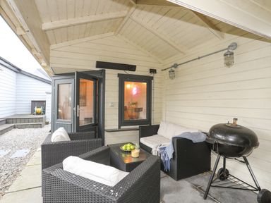 A covered patio area with black wicker chairs and a sofa around a glass table and a black barbecue grill at Laneton in Rhosneigr
