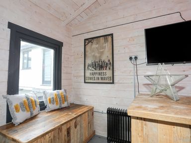 A small wooden room with a window bench with two cushions a wall poster a black radiator a wooden table a star-shaped glass decoration and a wall-mounted television at Laneton in Rhosneigr