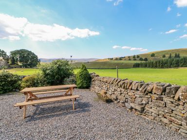 A wooden picnic table on a gravel area next to a stone wall with green fields and hills in the background at Field Barn in Alston