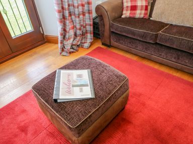 A brown ottoman with a welcome binder on a red rug next to a brown sofa and plaid curtains at Field Barn in Alston