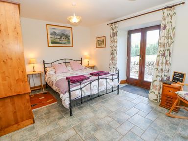 A bedroom with a double bed flanked by two bedside tables and lamps a wooden cabinet and rocking chair near double glass doors with floral curtains at Field Barn in Alston