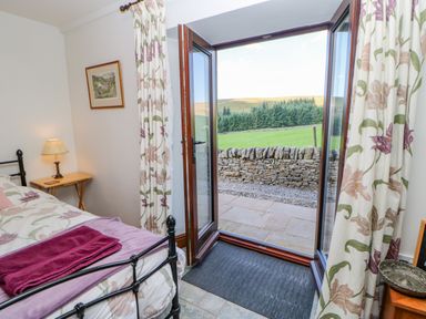 A bedroom with a bed and bedside table with lamp open doors leading to a stone patio and green fields at Field Barn in Alston