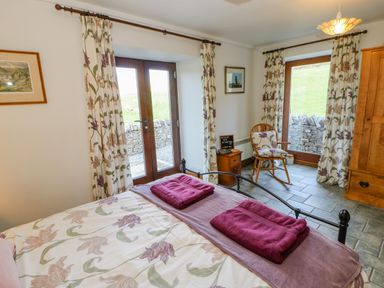 A bedroom with a bed with floral bedding two folded towels bedside table with a chair two large windows with floral curtains and a wooden wardrobe at Field Barn in Alston