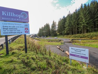 Two signs near a road and parking area with trees in the background at Killhope in Alston