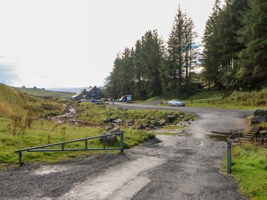A rural road with parked cars near a building and trees at Field Barn in Alston