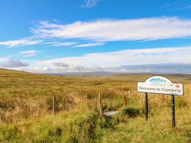 A landscape with grassy fields under a blue sky and a welcome to Cumbria sign at Field Barn in Alston