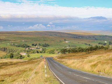 A countryside road leading through fields and hills with scattered houses and trees at Field Barn in Alston