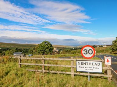 A rural road with a 30 mph speed limit sign and a Nenthead village sign next to a wooden fence in a countryside setting at Field Barn in Alston