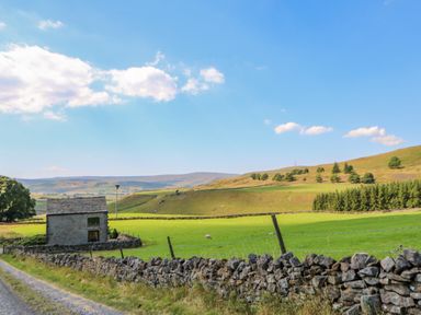 A stone barn near a gravel road and stone fence with green fields and hills at Field Barn in Alston