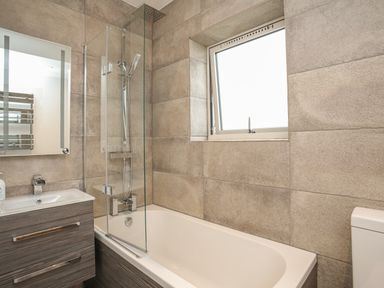 A bathroom with a bathtub shower combination a wooden vanity with sink a window and a toilet at Hafod Y Mor in Criccieth
