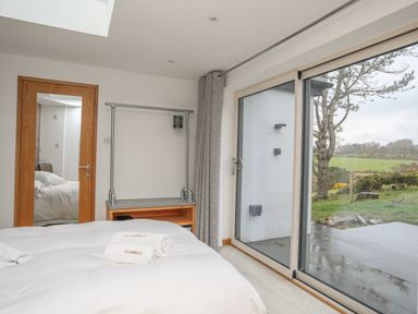 A bedroom with a bed, towels on the bed, a full-length mirror on a wooden door, a clothing rack, and large sliding glass doors showing a garden at Hafod Y Mor in Criccieth