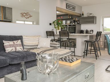 A living area with a sofa, armchairs, a coffee table with a chess set and binoculars, and a kitchen with bar stools at Hafod Y Mor in Criccieth