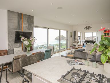 An open living space with a dining table, kitchen island with gas stove, chairs, flowers on tables, a television on a stone wall, large windows with a view of the sea, a hanging chair, and a foosball table at Hafod Y Mor in Criccieth