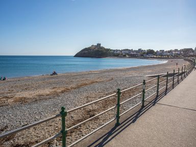 A pebble beach with a railing along a promenade and a hill with a castle in the background at Hafod Y Mor in Criccieth