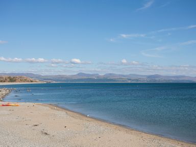 A beach with sand and pebbles beside a blue sea with hills in the background at Hafod Y Mor in Criccieth