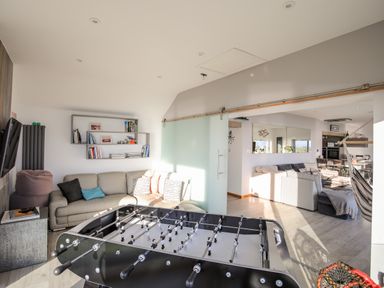 A room with a foosball table, a beige couch, a bookshelf, and a sliding door leading to a living area with sofas and a hammock at Hafod Y Mor in Criccieth