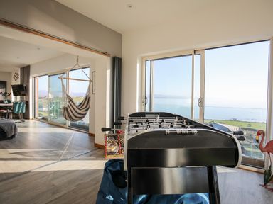 A room with a foosball table and a hanging hammock chair with large windows showing a sea view at Hafod Y Mor in Criccieth