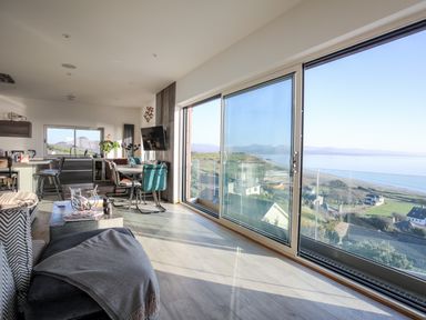 A living area with dining table chairs kitchen island and large sliding windows with a sea view at Hafod Y Mor in Criccieth