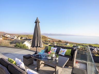 An outdoor terrace with rattan furniture a table with plants and blue glasses overlooking houses hills and a beach at Hafod Y Mor in Criccieth