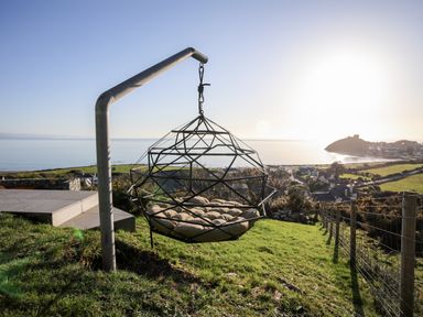 A metal hanging chair with cushions on a grassy hill overlooking a coastal town and sea at Hafod Y Mor in Criccieth