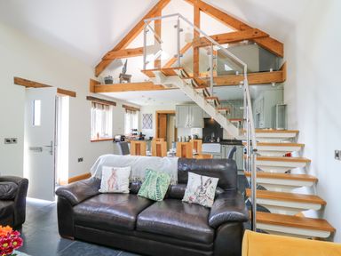 An open living room with a black leather sofa pillows a wooden dining table kitchen cabinets and a staircase with wooden steps and glass railing at Yr Hen Beudy in Capel Bangor