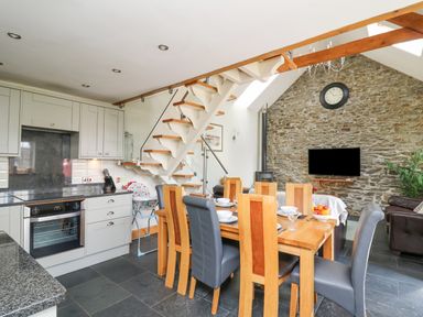 A kitchen and dining area with wooden table and chairs, staircase, stone wall with clock and mounted TV at Yr Hen Beudy in Capel Bangor