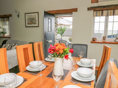 A dining table set with dishes glasses and a flower vase near an open door and window at Yr Hen Beudy in Capel Bangor