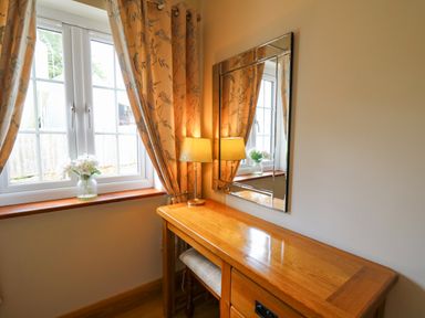 A wooden dressing table with a lamp and a mirror next to a window with curtains and a vase of flowers at Yr Hen Beudy in Capel Bangor