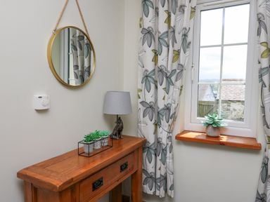 A wooden table with two small potted plants and a dog figurine lamp under a round mirror next to a window with floral curtains at Yr Hen Beudy in Capel Bangor