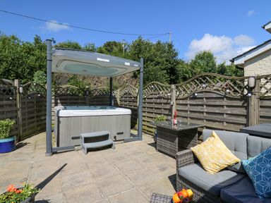 An outdoor patio with a hot tub under a canopy a wicker table with a bottle and glasses and a cushioned sofa with pillows at Yr Hen Beudy in Capel Bangor