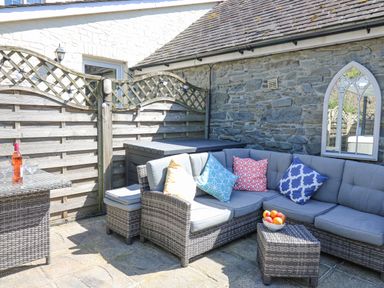 An outdoor patio with wicker corner sofa with cushions a small wicker table with a bowl of fruit a glass table with wine and glasses and a wooden fence at Yr Hen Beudy in Capel Bangor