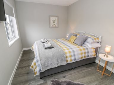 A bedroom with a bed with checked bedding and pillows a side table with a lamp and a window at Cartref in Trawsfynydd