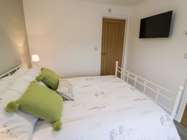A bedroom with a bed with white and floral bedding green pillows a wooden door a wall lamp and a wall-mounted TV at Cartref in Trawsfynydd