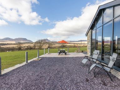 An outdoor patio with two reclining chairs and a picnic table with an orange umbrella overlooking a grassy field and hills at Cartref in Trawsfynydd