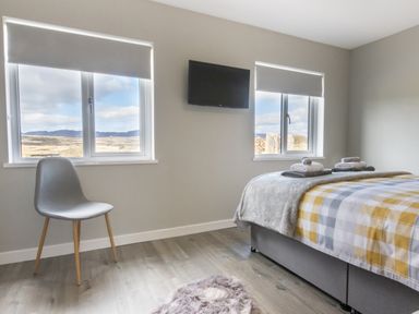 A bedroom with a bed covered in a checkered duvet and folded towels a gray chair two windows and a mounted TV at Cartref in Trawsfynydd