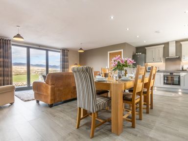 A dining table with chairs and flowers next to a kitchen and a living area with sofas and large windows at Cartref in Trawsfynydd