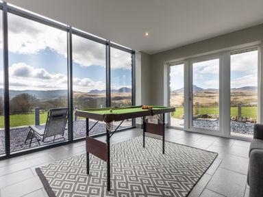 A recreational room with a small pool table on a patterned rug and large windows showing an outdoor landscape at Cartref in Trawsfynydd