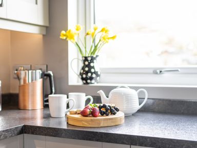 A kitchen countertop with a teapot mugs fruit and a vase of flowers on the windowsill at Cartref in Trawsfynydd
