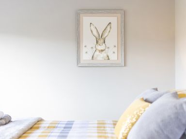 A bed with gray and yellow pillows and a checkered blanket under a framed rabbit painting on a wall at Cartref in Trawsfynydd