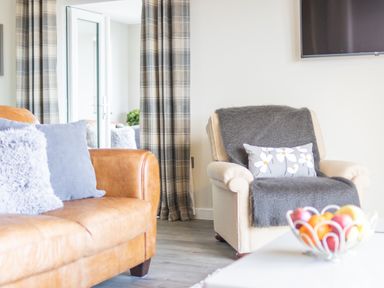 A living room with a leather sofa and armchair with cushions and a bowl of fruit on a table at Cartref in Trawsfynydd