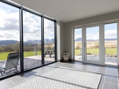 A room with large glass windows and doors showing a patio table and chairs and a mountain view at Cartref in Trawsfynydd