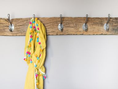 A wooden coat rack with five metal hooks and a yellow scarf with colorful pom poms hanging on one hook at Cartref in Trawsfynydd