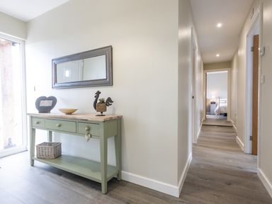 A hallway with a green console table with drawers a basket and decorative items a mirror on the wall and a bedroom visible at the end of the hallway at Cartref in Trawsfynydd