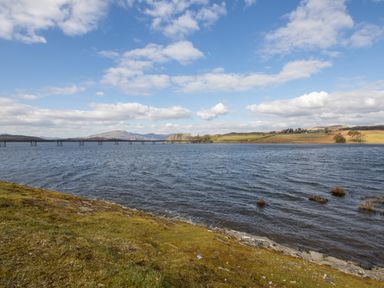 A lake with a long bridge across it and grassy hills in the background at Cartref in Trawsfynydd