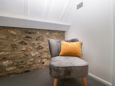 A grey upholstered chair with wooden legs and a yellow cushion in a corner with stone and white walls at The Linhay in Chardstock
