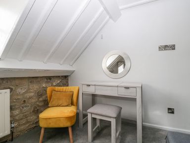 A small dressing table with a round mirror a padded stool and a yellow chair with a cushion in an attic room with slanted ceiling and stone wall at The Linhay in Chardstock