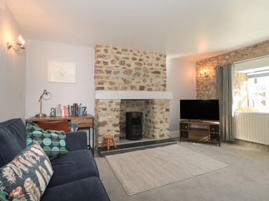 A living room with a sofa a desk with books a stone fireplace a TV on a stand and a window with curtains at The Linhay in Chardstock