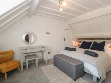 A bedroom with a bed, gray bench, vanity table, round mirror, yellow chair, and slanted ceiling at The Linhay in Chardstock