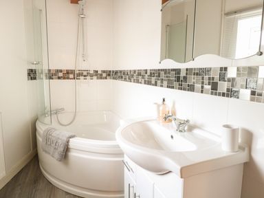 A bathroom with a corner bathtub shower and sink with mosaic tile border at Compass Cottage in Bodham near Sheringham